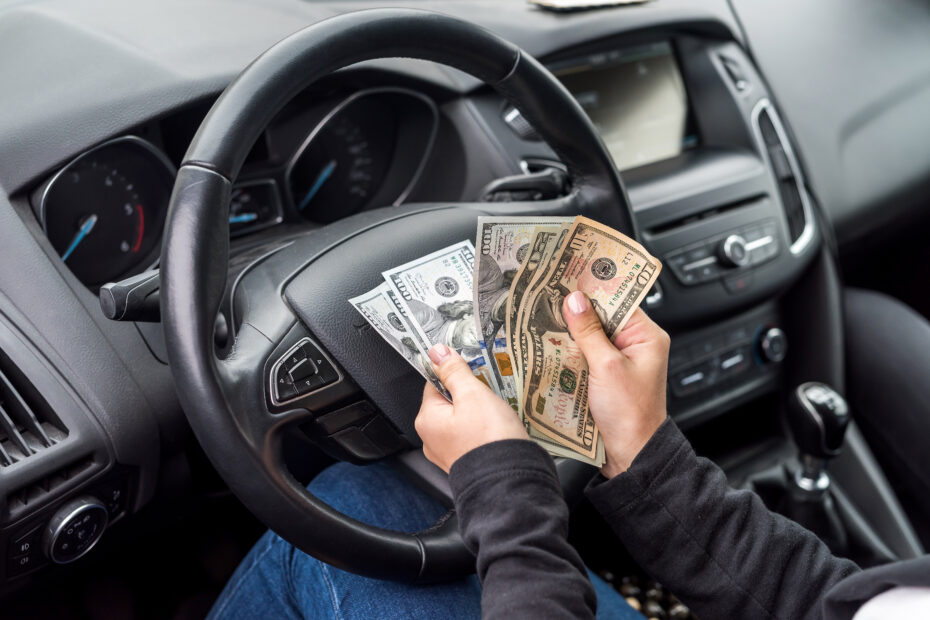 Person holding a fan of U.S. dollar bills over a car’s steering wheel.
