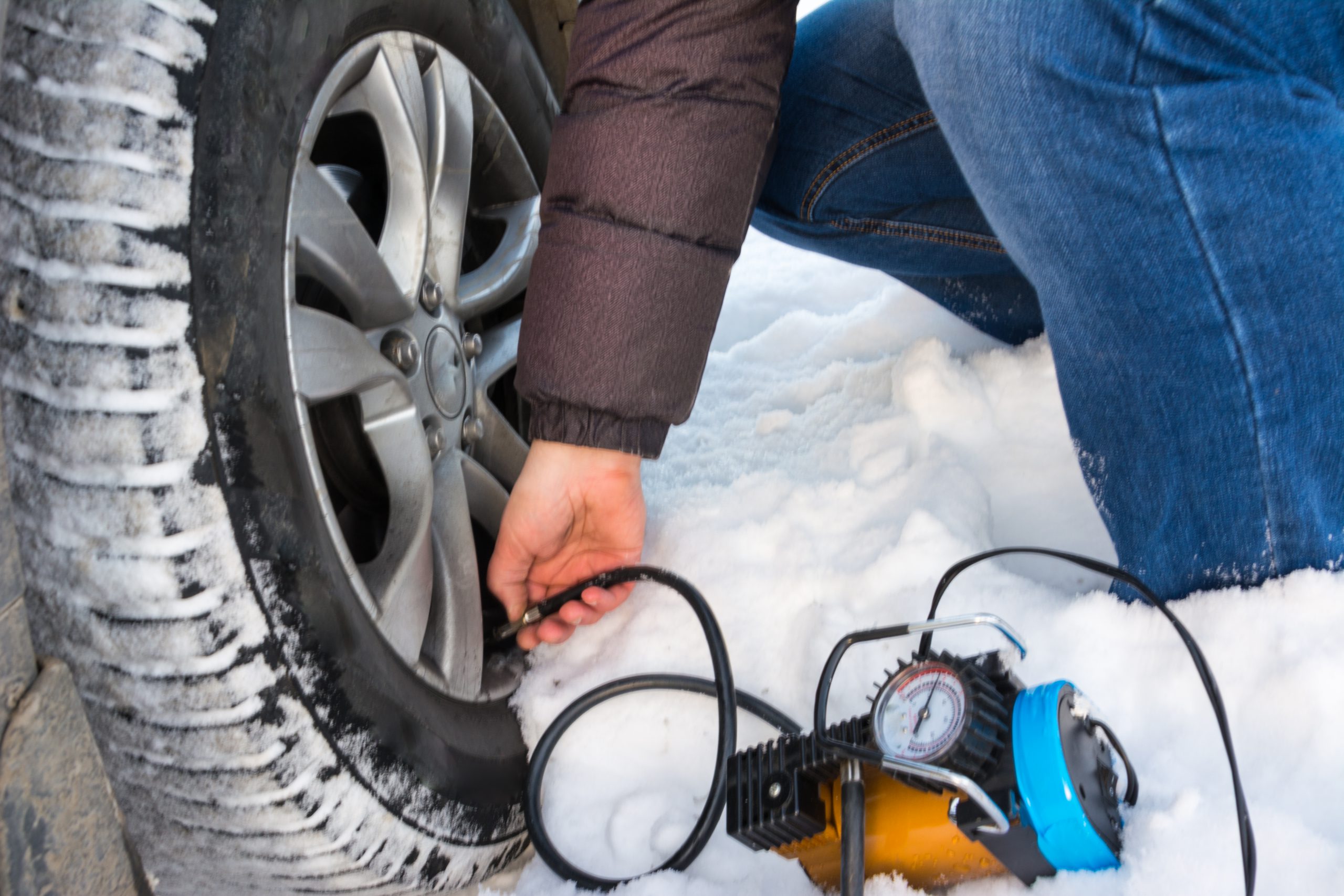 winter-tire-inflation-air-compressor A person filling air into a car tire with a portable air compressor on a snowy day.