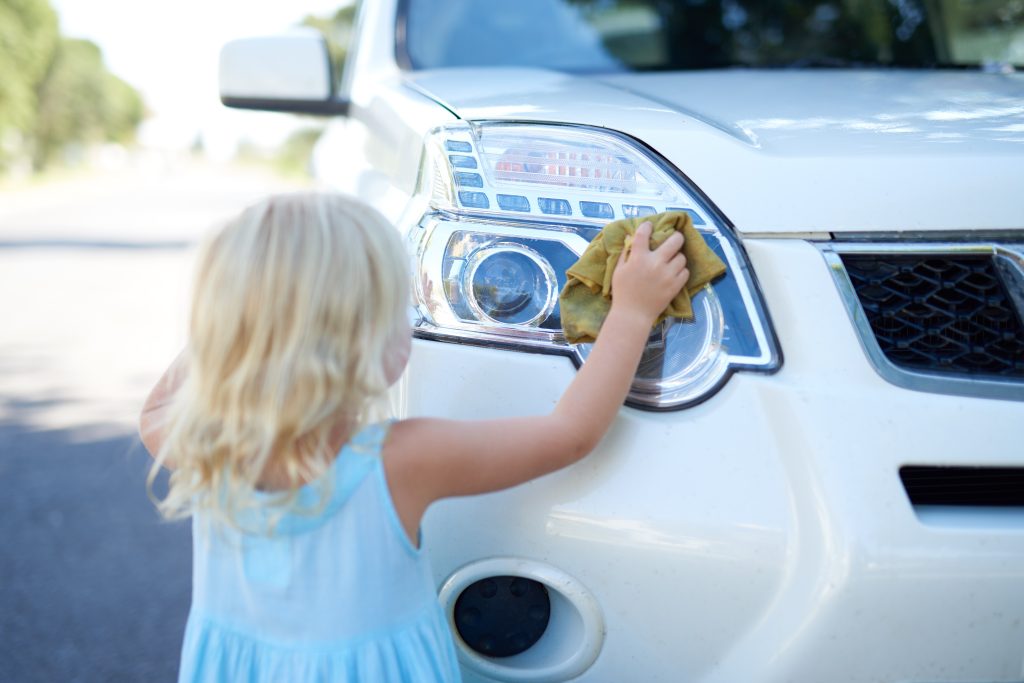 A young girl in a blue dress helping to wash the headlight of a white car outdoors.