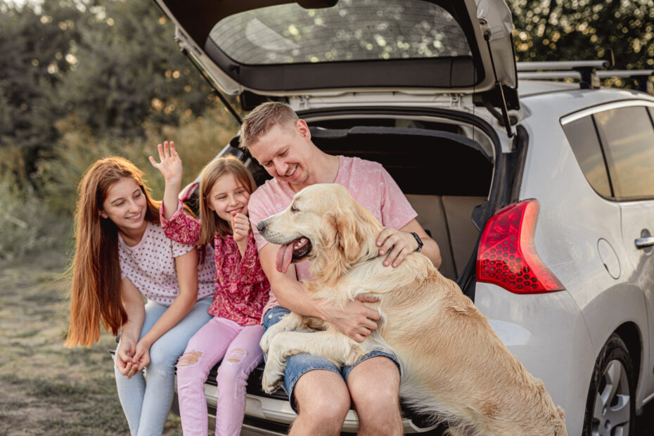A father and his two daughters smiling and sitting with their golden retriever in the open trunk of a car.