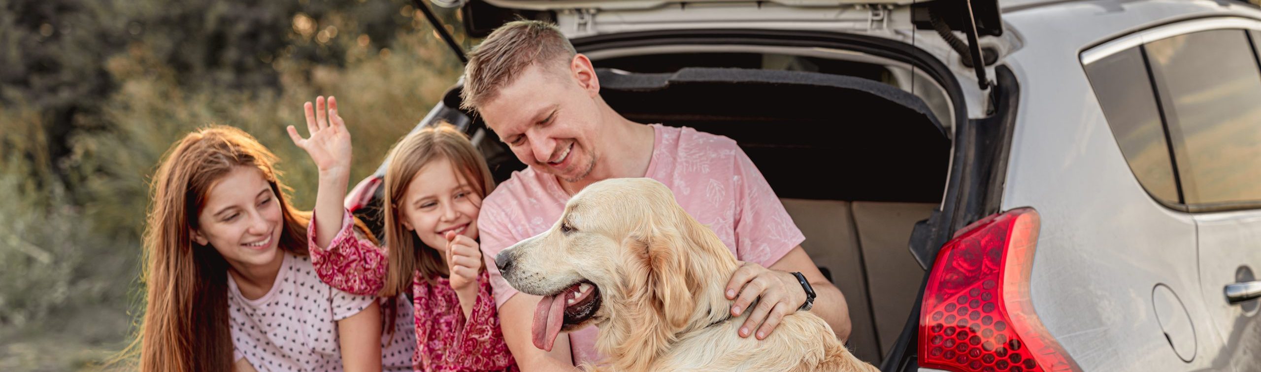 A father with his daughters and dog sitting in an open car trunk outdoors.