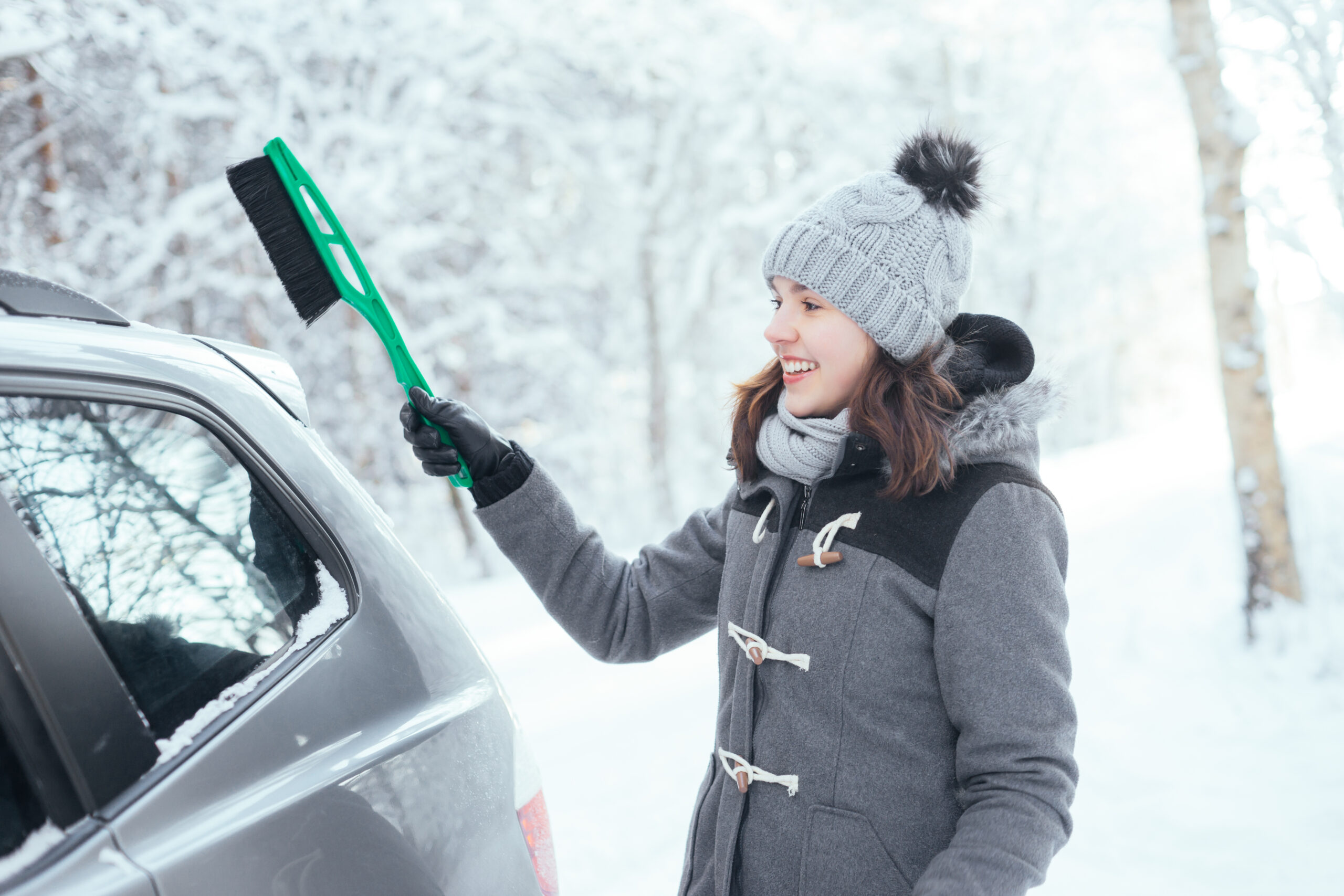 Smiling woman brushing snow off her car with a snow brush during winter.