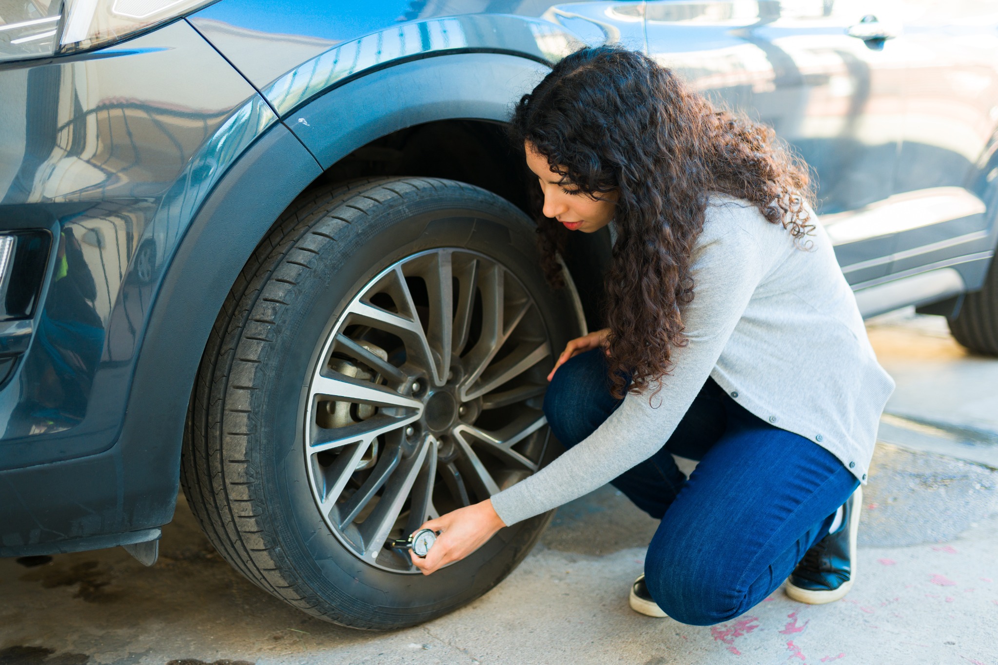 A woman crouches beside her car, checking the tire pressure with a gauge to ensure safe spring driving.