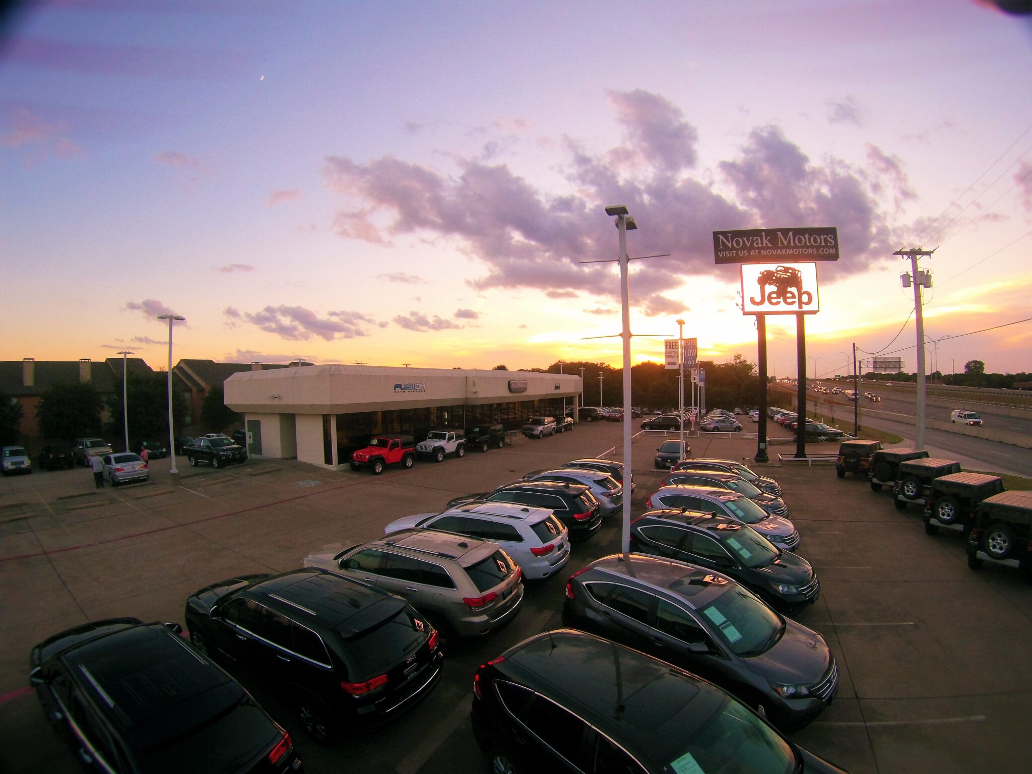Sunset view of Novak Motors dealership in Bedford, Texas, showcasing parked vehicles and a Jeep sign, celebrating Fusion Auto Finance’s 2012 milestone of 20,000 vehicles leased through CU Xpress Lease.